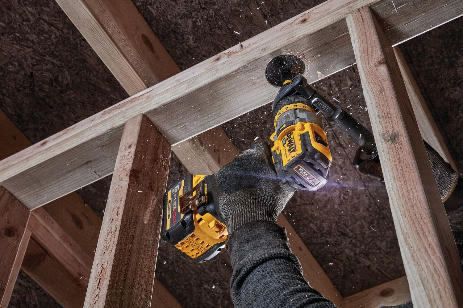 Una persona usando guantes emplea un taladro inalámbrico DEWALT para hacer un agujero en una viga de madera durante trabajos de construcción. Se ven virutas de madera mientras el taladro está en funcionamiento.