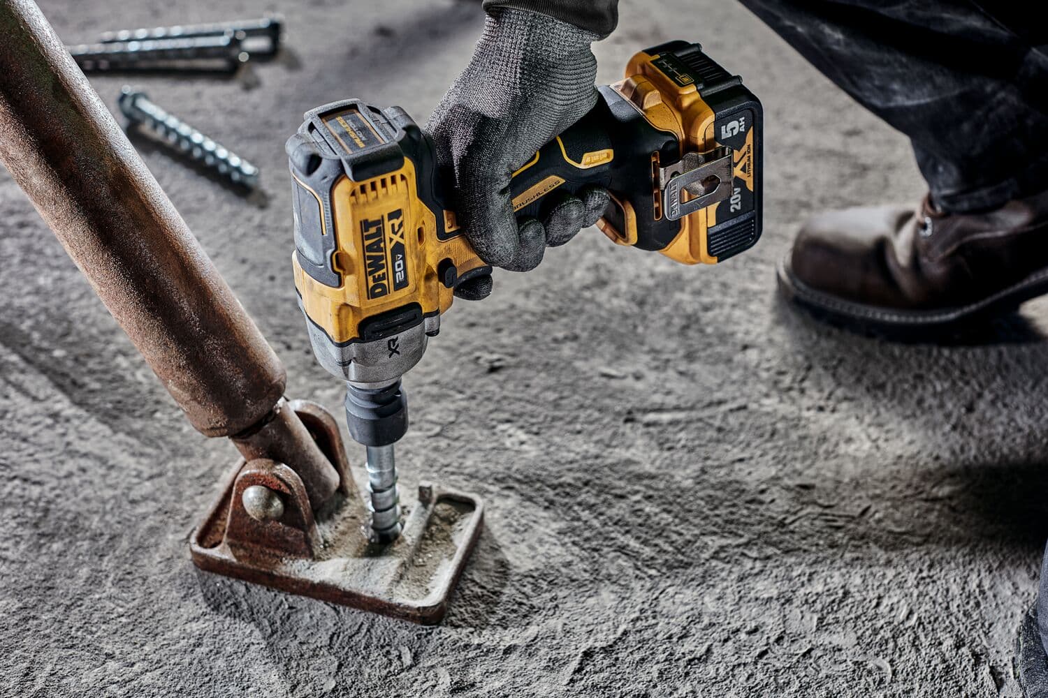 Vista en primer plano de una mano enguantada usando una llave de impacto inalámbrica DEWALT 20V (modelo DCF892B) para asegurar una base metálica a un piso de concreto. La herramienta es amarilla y negra, y el trabajo se realiza sobre una superficie industrial áspera.