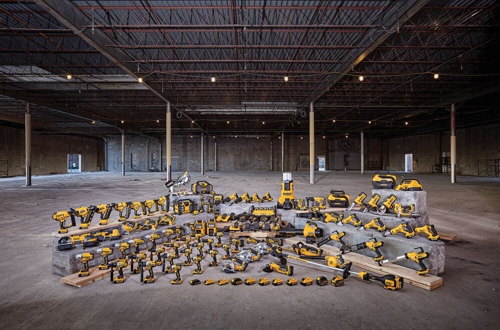 Una gran variedad de herramientas eléctricas inalámbricas y accesorios DEWALT amarillos y negros exhibidos sobre bloques de concreto y madera dentro de un almacén vacío.