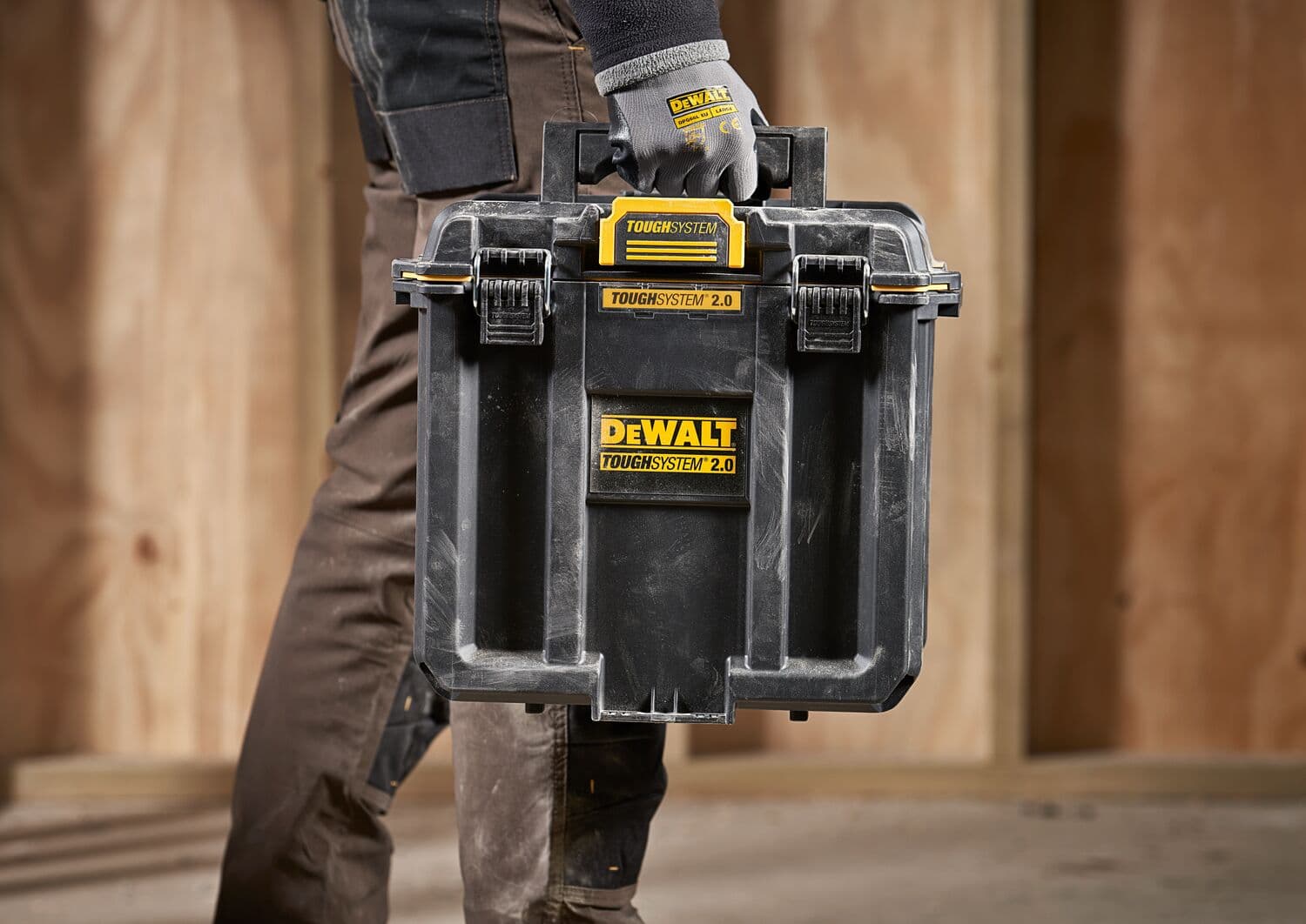 Una persona con guantes de trabajo transporta una caja de almacenamiento negra DEWALT TOUGHSYSTEM 2.0 con etiquetas amarillas, diseñada para organizar y transportar herramientas, en un entorno de construcción con paredes de madera.