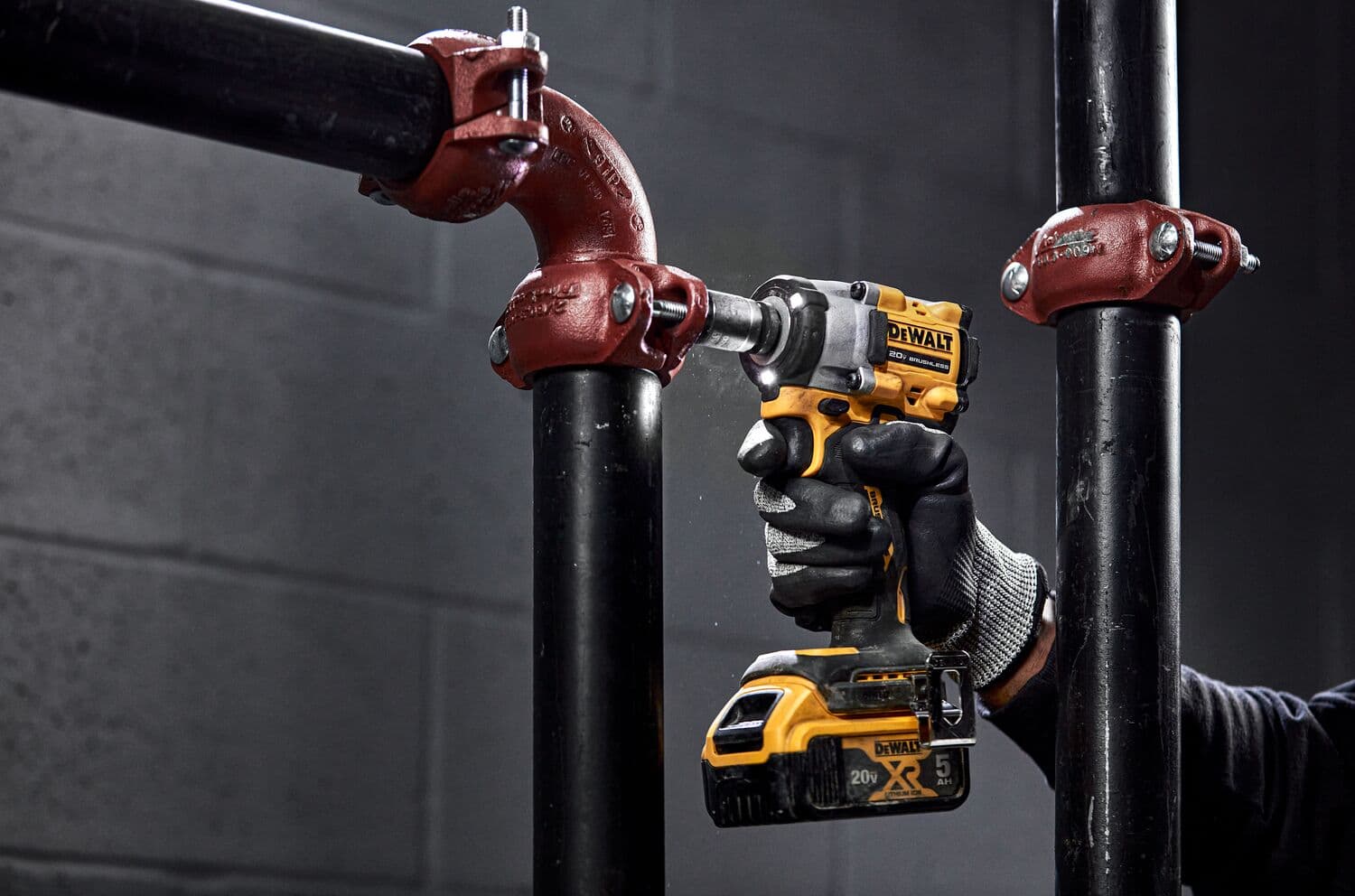 A person wearing gloves uses a DEWALT cordless impact wrench to tighten a bolt on a black metal pipe with red fittings in an industrial setting.