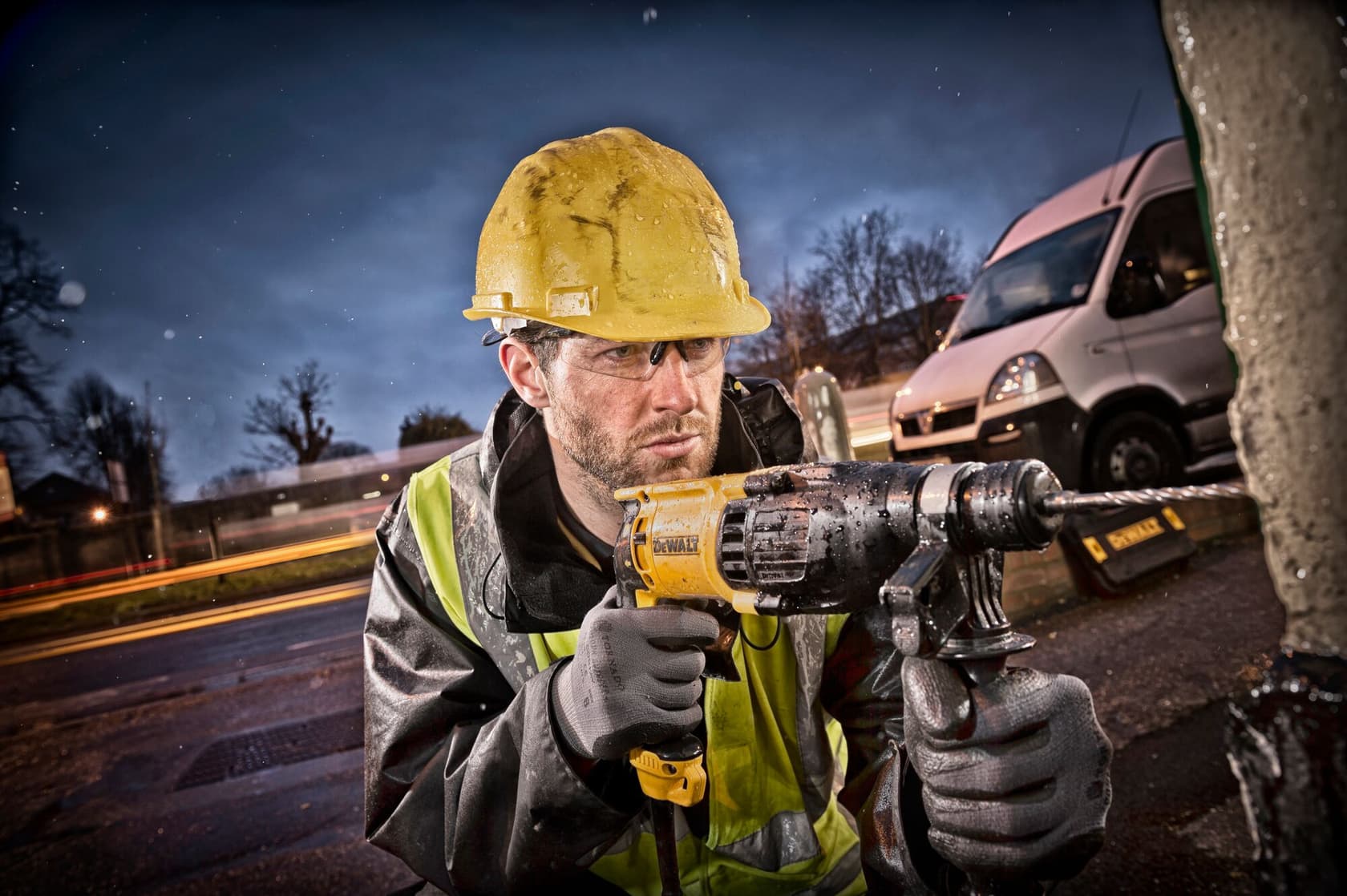 Una persona con casco amarillo y chaleco reflectante utiliza un taladro eléctrico DEWALT al aire libre, con una furgoneta y luces borrosas visibles en el fondo. El rostro de la persona está oculto.