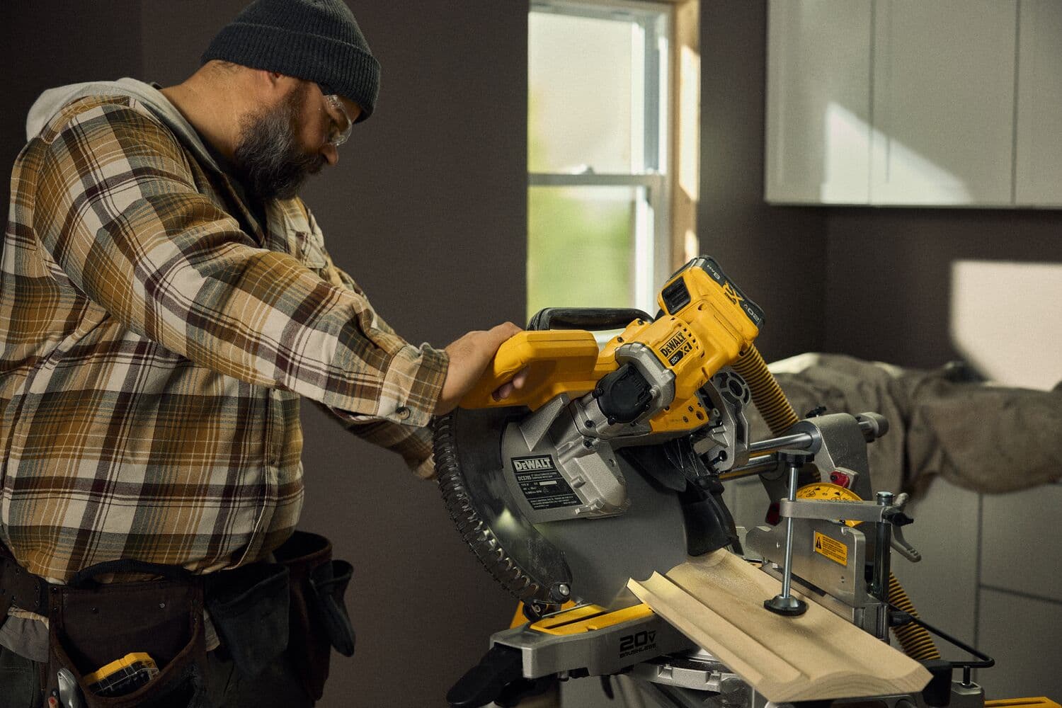 Una persona opera una sierra de inglete inalámbrica DEWALT DCS785B para cortar molduras de madera en un taller interior bien iluminado. El trabajador lleva una camisa de cuadros, un cinturón de herramientas y un gorro. La sierra está sobre un banco de trabajo, y la habitación tiene gabinetes y una ventana.